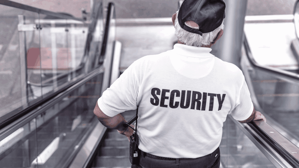 Image of a middle-aged man wearing a T-shirt with the text "Security" on the back walking down an escalator as a representation of store security systems.