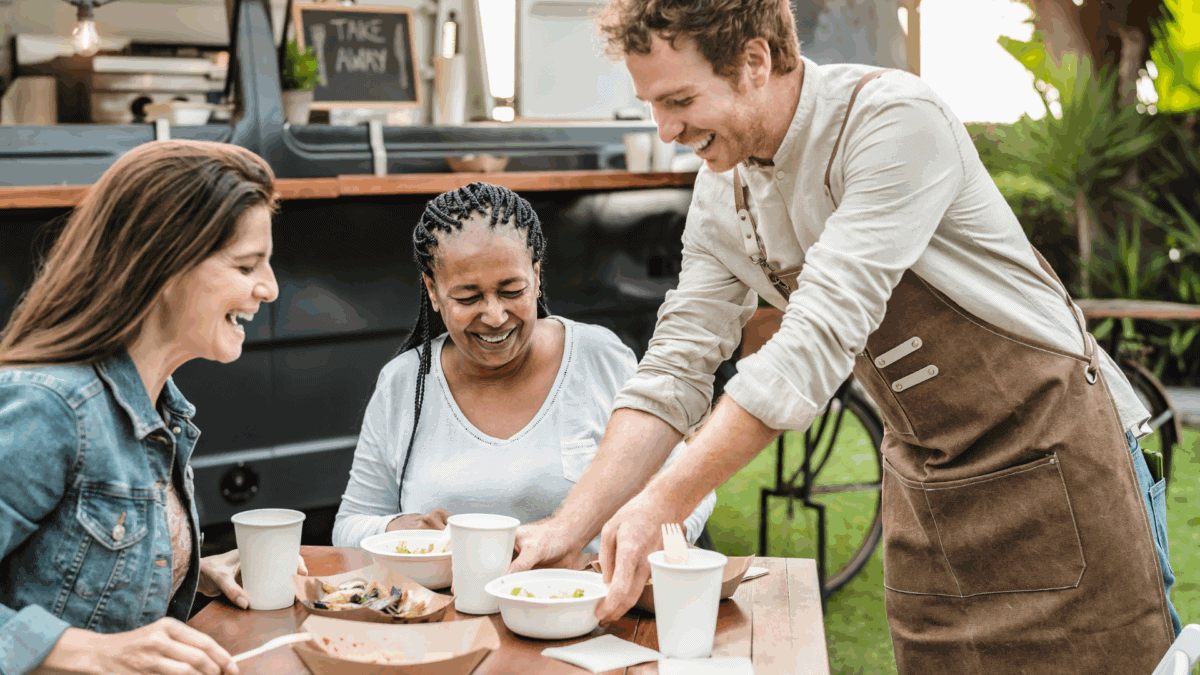 A server happily serving food to two female customers outside a food truck. A great example of QSR customer experience.