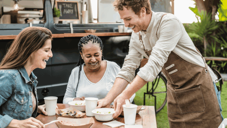 A server happily serving food to two female customers outside a food truck. A great example of QSR customer experience.