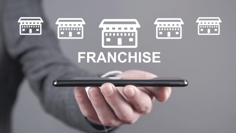 Close-up photograph of a man's hand holding a cell phone with white icon illustrations of five stores and, below, the word "Franchise" written as a representation of retail franchise opportunities.