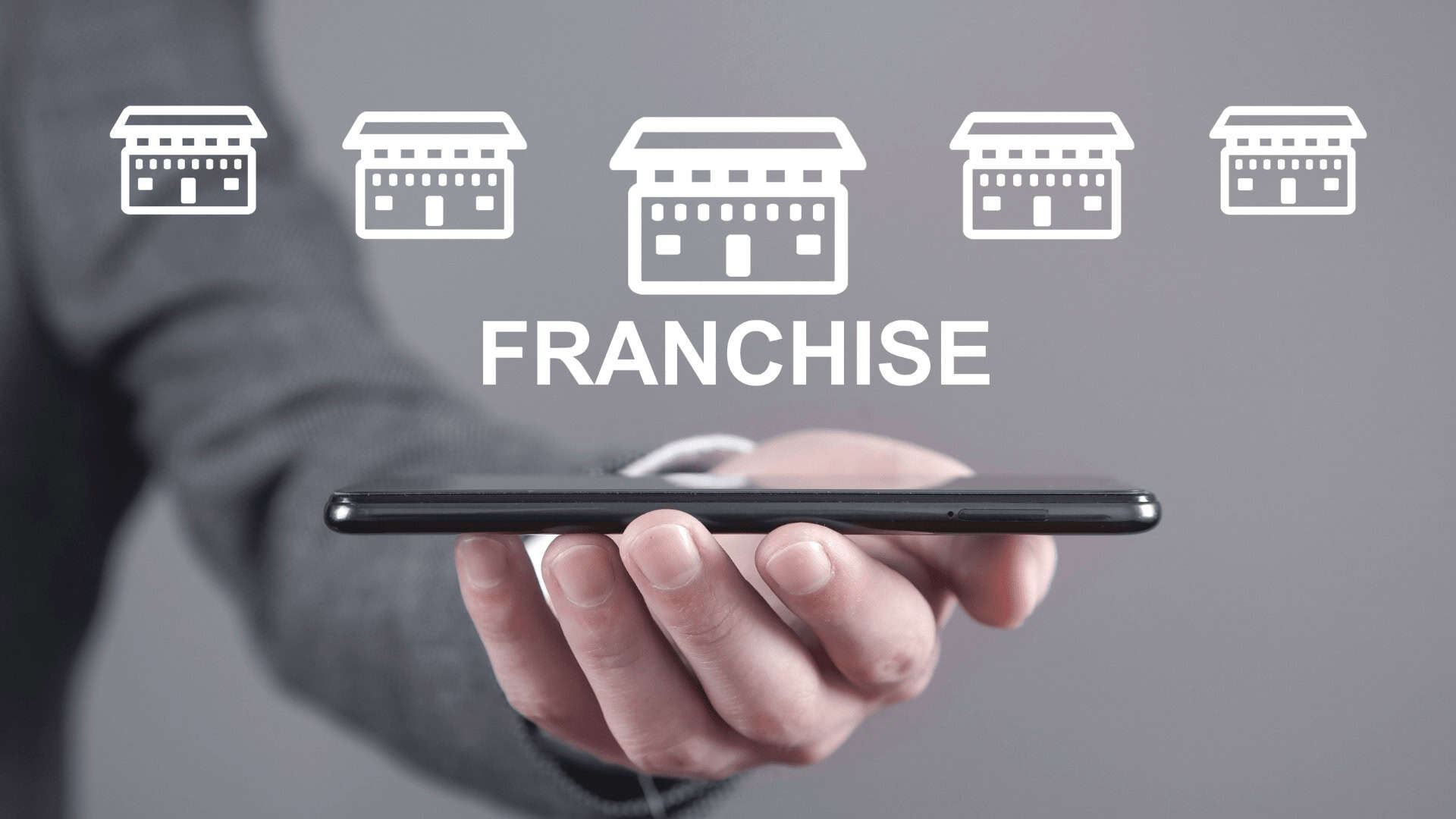 Close-up photograph of a man's hand holding a cell phone with white icon illustrations of five stores and, below, the word "Franchise" written as a representation of retail franchise opportunities.