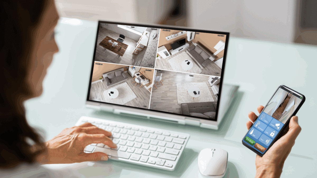 A person monitors a home security system via a desktop screen showing four live camera feeds, while holding a smartphone with a surveillance app, visually explaining how does a hotspot camera work through real-time remote access and control.