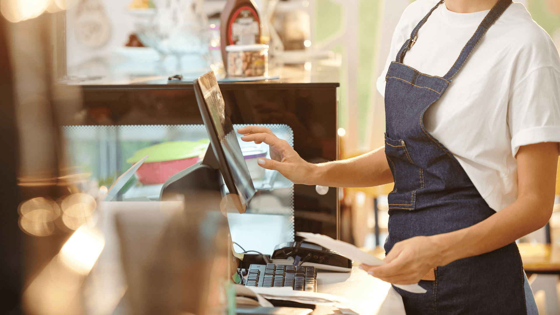 A retail worker in an apron operates a point-of-sale system while holding receipts, representing frontline roles in safeguarding store operations—an ideal visual for discussing loss prevention vs asset protection.