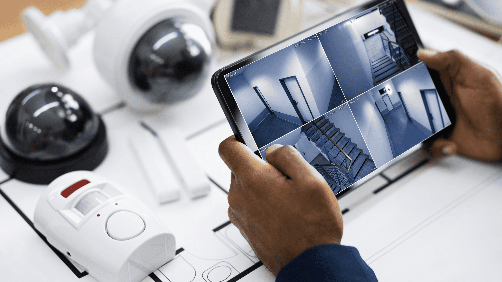 A person holds a tablet displaying multiple live camera feeds from a building interior, surrounded by various security devices, illustrating the use and functionality of a CCTV monitor in surveillance systems.