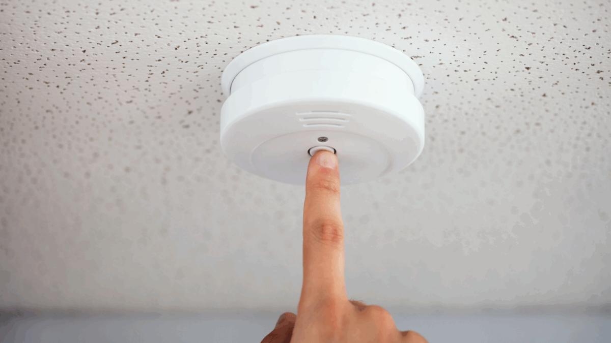A hand reaches up to press the button on a ceiling-mounted motion sensor device, representing a key component of motion detector camera security systems.