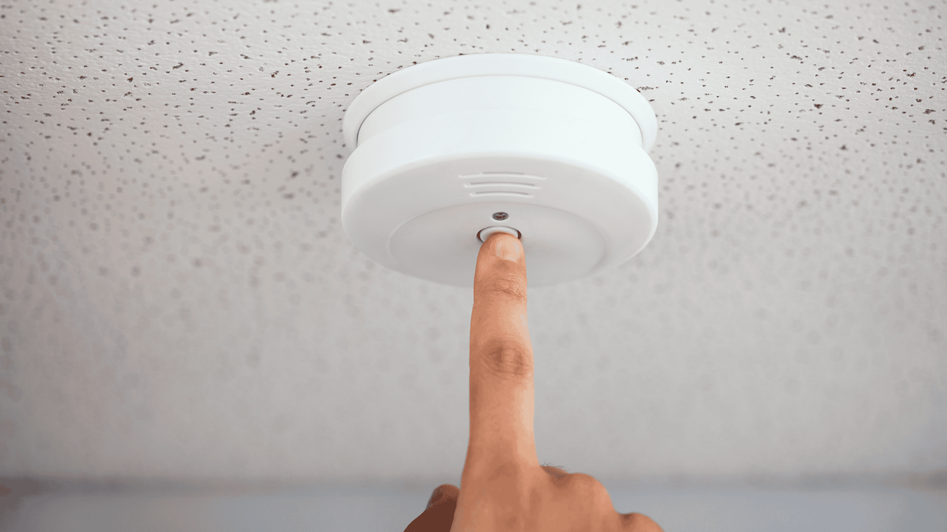 A hand reaches up to press the button on a ceiling-mounted motion sensor device, representing a key component of motion detector camera security systems.