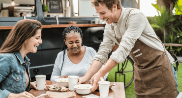 A server happily serving food to two female customers outside a food truck. A great example of QSR customer experience.