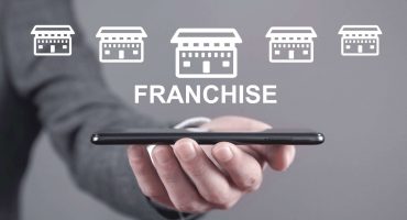 Close-up photograph of a man's hand holding a cell phone with white icon illustrations of five stores and, below, the word "Franchise" written as a representation of retail franchise opportunities.