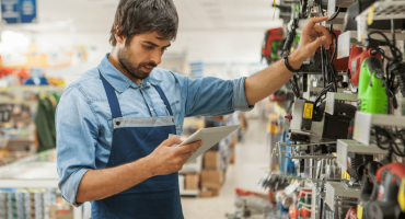 A retail employee in a blue apron checks inventory using a tablet while inspecting products on a store shelf, illustrating a key task in the morning checklist for opening a retail store.