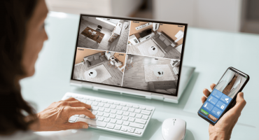 A person monitors a home security system via a desktop screen showing four live camera feeds, while holding a smartphone with a surveillance app, visually explaining how does a hotspot camera work through real-time remote access and control.